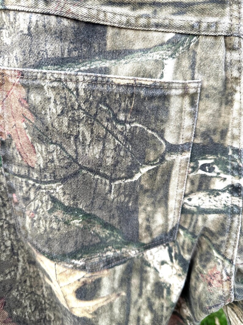 May include: Close-up of camouflage-patterned denim fabric, featuring shades of green, brown, and gray. The image highlights a back pocket and stitching details, showcasing the texture and design of the material. The overall theme is outdoor apparel.