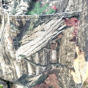 May include: Close-up of a pair of camouflage pants. The pants feature a detailed pattern of brown, green, and tan, mimicking a forest environment. The back pocket is visible, with stitching details.