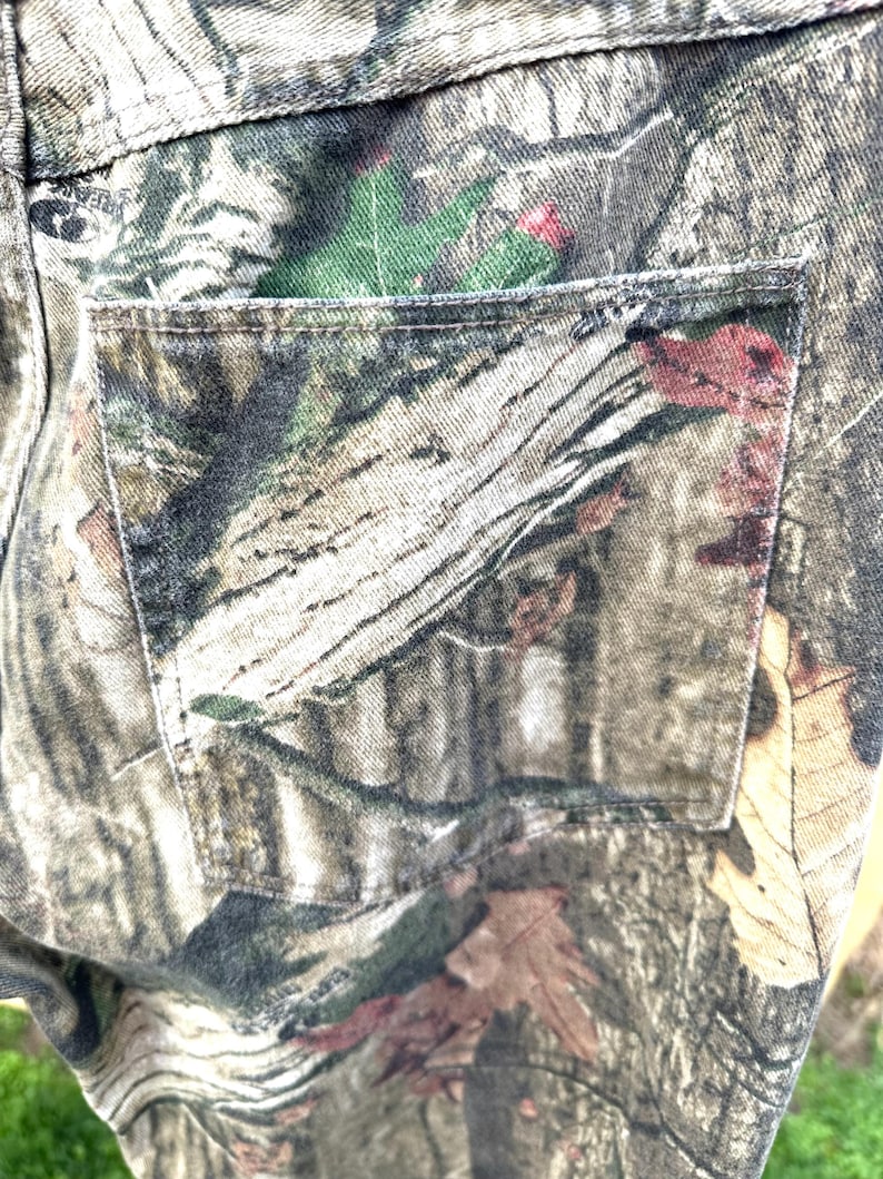 May include: Close-up of a pair of camouflage pants. The pants feature a detailed pattern of brown, green, and tan, mimicking a forest environment. The back pocket is visible, with stitching details.