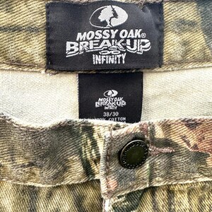 May include: Close-up of camouflage pants with the "MOSSY OAK BREAK-UP INFINITY" logo on a black tag. The pants have a button closure and a size tag that reads "38/30". The fabric is 100% cotton.