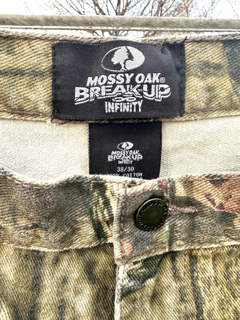 May include: Close-up of camouflage pants with the "MOSSY OAK BREAK-UP INFINITY" logo on a black tag. The pants have a button closure and a size tag that reads "38/30". The fabric is 100% cotton.