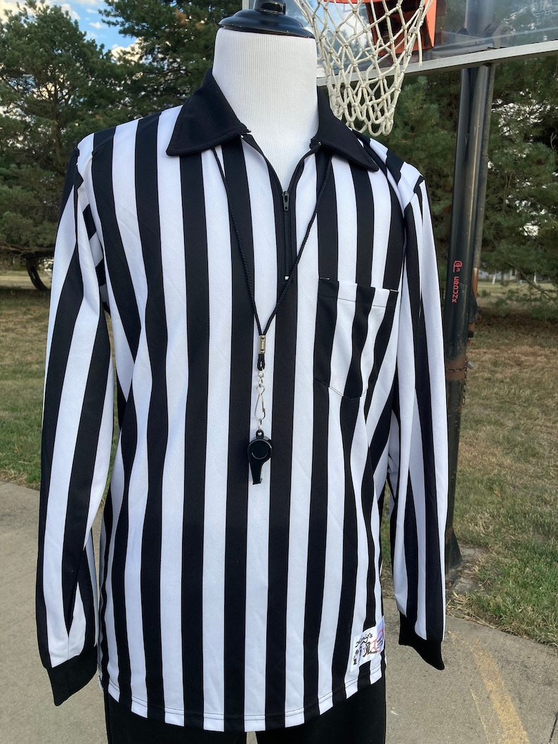 May include: Black and white striped referee shirt with a zipper and a whistle on a black lanyard. The shirt has a pocket on the left side.
