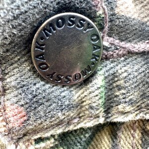 May include: Close-up of a metal button with the words "OAK-MOSSY" engraved around the edge. The button is attached to a camouflage-patterned fabric in shades of green, brown, and gray. A thread is visible near the button.