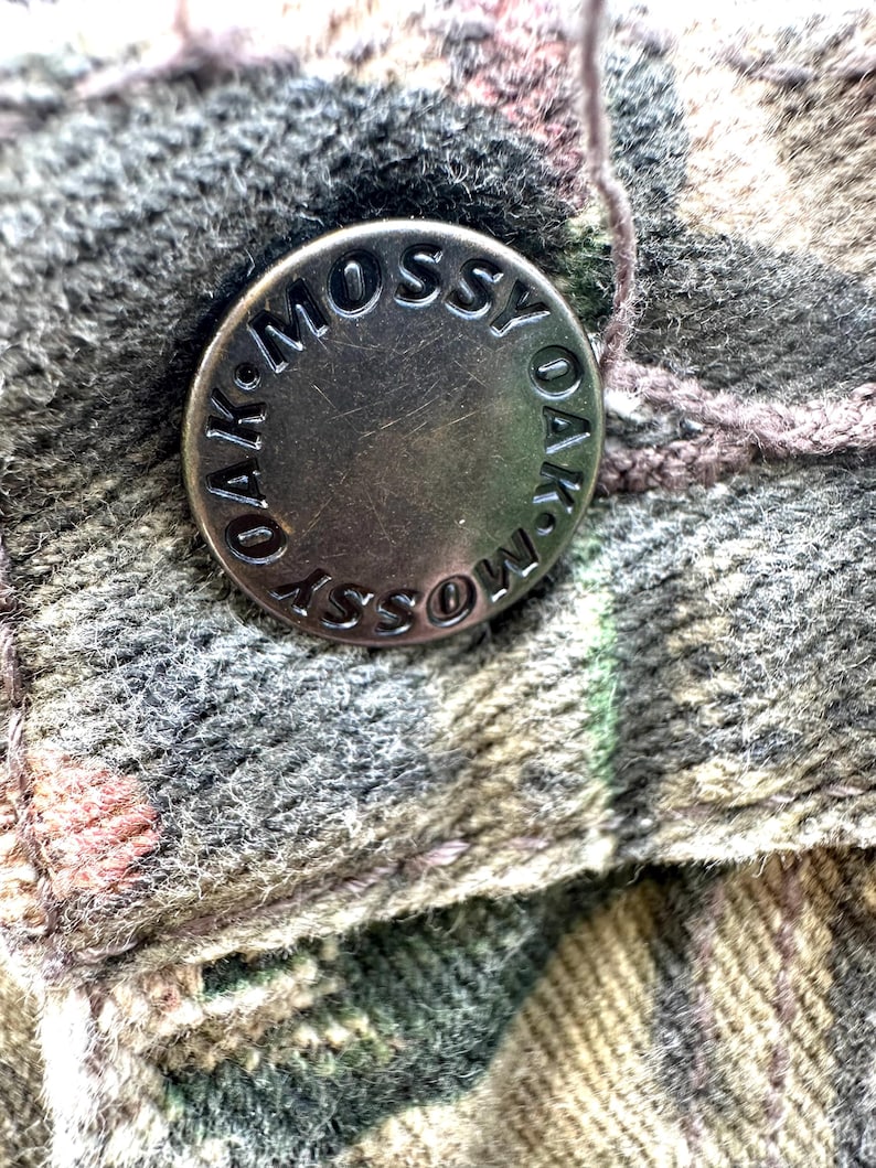 May include: Close-up of a metal button with the words "OAK-MOSSY" engraved around the edge. The button is attached to a camouflage-patterned fabric in shades of green, brown, and gray. A thread is visible near the button.