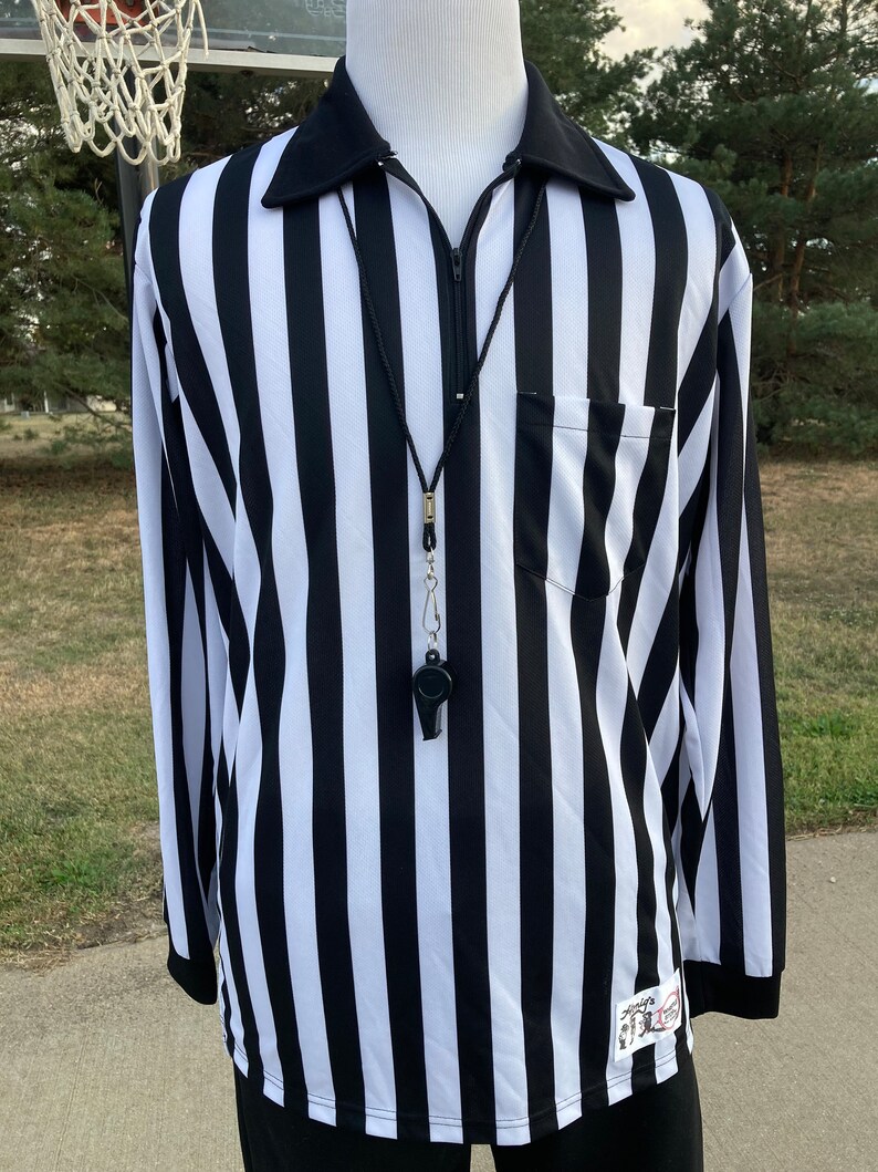 May include: Black and white striped referee shirt with a zipper and a whistle on a black lanyard. The shirt has a small pocket on the left chest and a logo on the right sleeve.