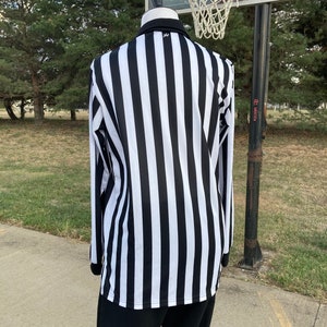 May include: A black and white striped referee shirt with long sleeves. The shirt is on a mannequin and is standing in front of a basketball hoop.