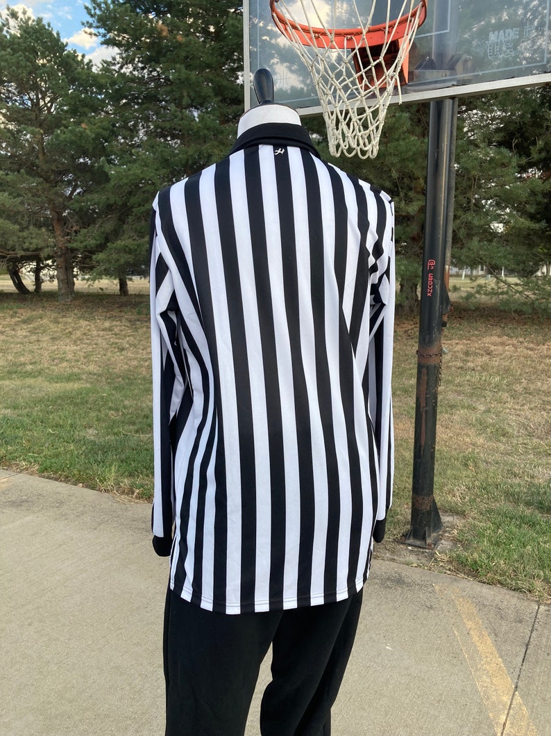May include: A black and white striped referee shirt with long sleeves. The shirt is on a mannequin and is standing in front of a basketball hoop.