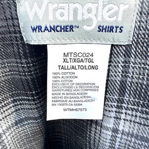 May include: Close-up of a gray and white plaid Wrangler shirt label. The label reads "Wrangler WRANCHER SHIRTS" and includes details about the shirt's material and origin. The shirt is made in Bangladesh.