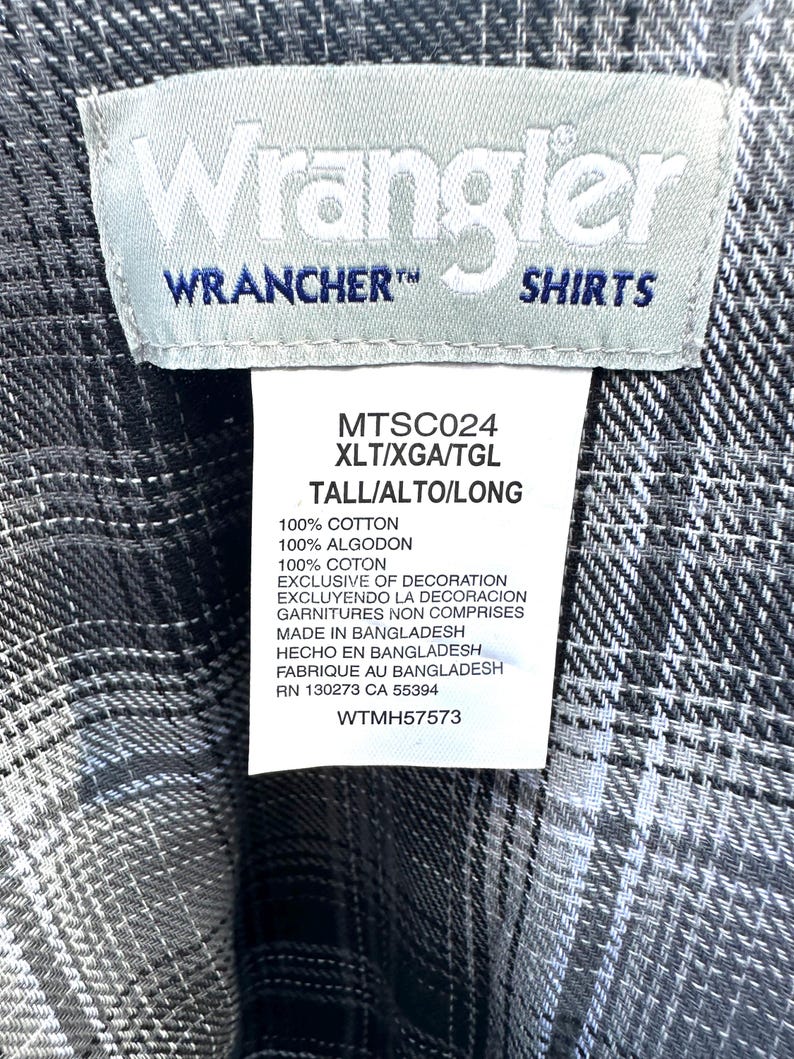 May include: Close-up of a gray and white plaid Wrangler shirt label. The label reads "Wrangler WRANCHER SHIRTS" and includes details about the shirt's material and origin. The shirt is made in Bangladesh.