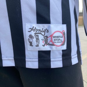 May include: A black and white striped referee shirt with a patch that says "Honig's Whistle Stop Inc. Ann Arbor, MI"