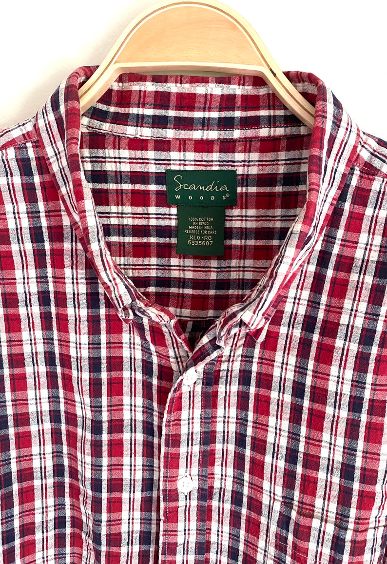 May include: A red, white, and blue plaid button-down shirt with a Scandia Woods label. The label says "100% Cotton FN 81700 Made in India Reverse for Care XL-RG 5335607".