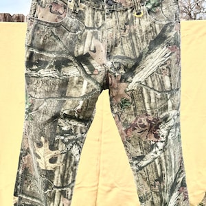 May include: Camouflage-patterned jeans hanging on a clothesline. The pants feature a mix of green, brown, and tan hues, creating a realistic woodland camouflage design. The jeans have a button and zipper closure, with pockets and belt loops.