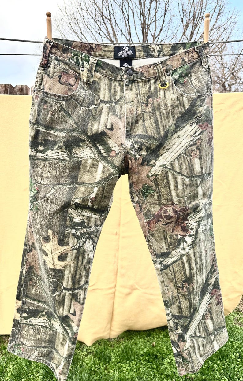 May include: Camouflage-patterned jeans hanging on a clothesline. The pants feature a mix of green, brown, and tan hues, creating a realistic woodland camouflage design. The jeans have a button and zipper closure, with pockets and belt loops.