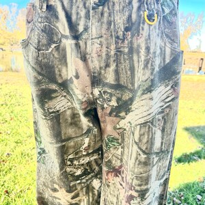 May include: Camouflage-patterned pants in shades of green, brown, and tan. The pants have a button and zipper closure, belt loops, and pockets. A gold-colored ring is attached to the belt loop. The pants are made of a durable fabric.