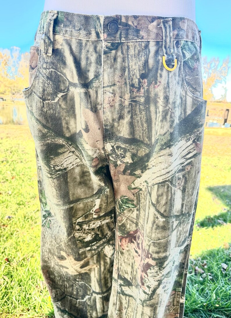 May include: Camouflage-patterned pants in shades of green, brown, and tan. The pants have a button and zipper closure, belt loops, and pockets. A gold-colored ring is attached to the belt loop. The pants are made of a durable fabric.