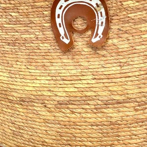 May include: Close-up of a straw hat with a brown leather horseshoe emblem. The hat has a woven texture and a brown leather band. The horseshoe is white with a brown leather backing.