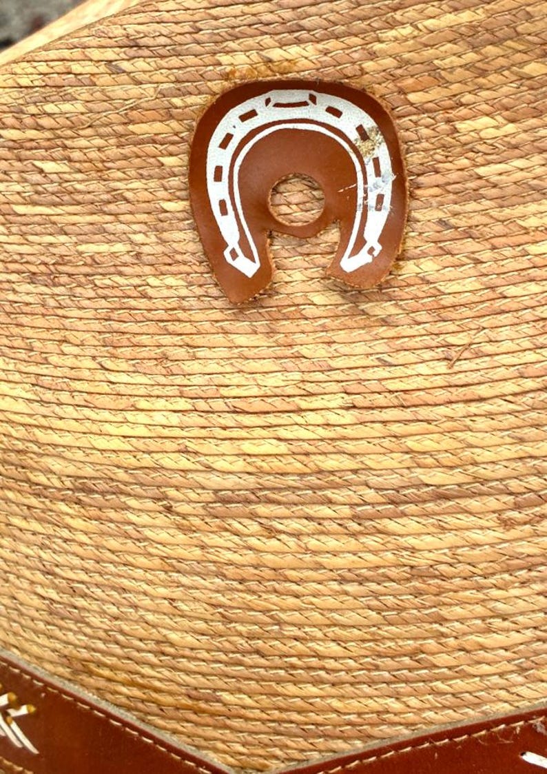 May include: Close-up of a straw hat with a brown leather horseshoe emblem. The hat has a woven texture and a brown leather band. The horseshoe is white with a brown leather backing.