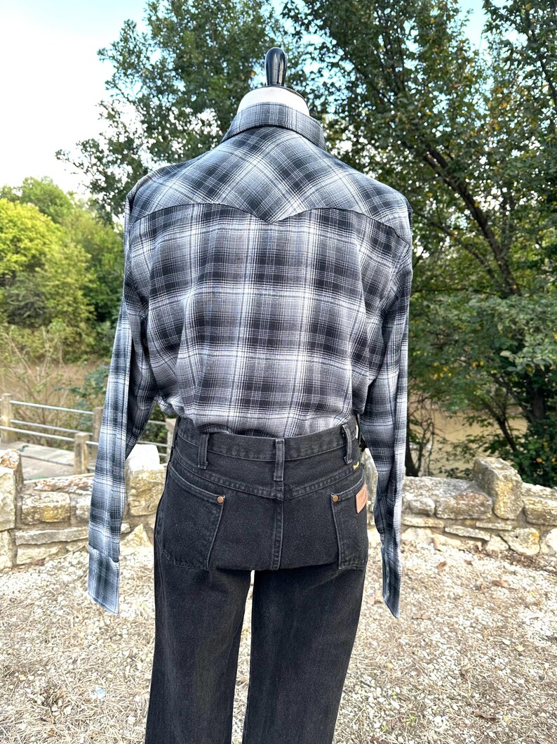 May include: A black and white plaid long-sleeve shirt and black denim jeans. The shirt has a classic western style with a pointed yoke and buttoned cuffs. The jeans are a dark wash with a high waist and a straight leg.