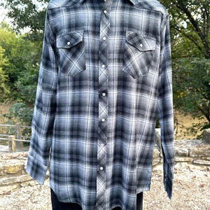 May include: A long-sleeved plaid shirt in shades of black, white, and gray. The shirt features a button-down front, two chest pockets, and a classic collar. The shirt is displayed on a mannequin.