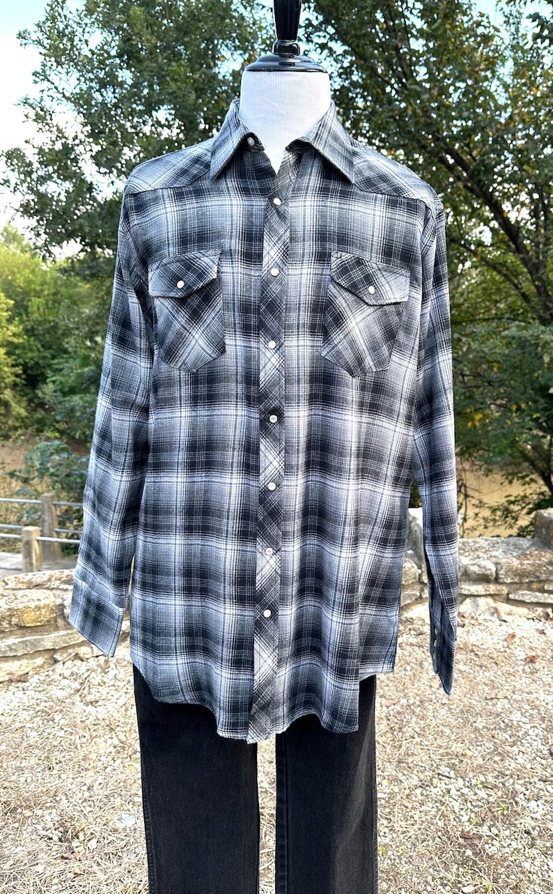 May include: A long-sleeved plaid shirt in shades of black, white, and gray. The shirt features a button-down front, two chest pockets, and a classic collar. The shirt is displayed on a mannequin.