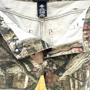 May include: Camouflage-patterned pants with a button and zipper closure. The pants feature the "Mossy Oak Break-Up Infinity" logo on the inside waistband. The fabric has a mix of green, brown, and tan colors.