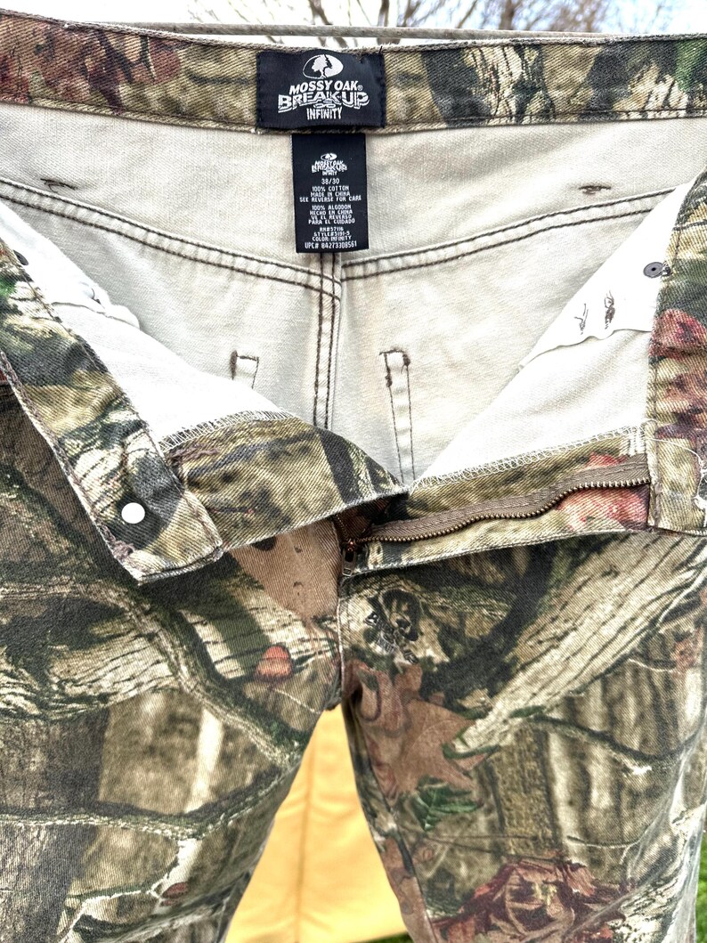 May include: Camouflage-patterned pants with a button and zipper closure. The pants feature the "Mossy Oak Break-Up Infinity" logo on the inside waistband. The fabric has a mix of green, brown, and tan colors.