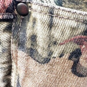 May include: Close-up of camouflage-patterned fabric, featuring shades of brown, green, and black. A dark button is visible on the pocket. The fabric appears to be denim or a similar sturdy material, with visible stitching.