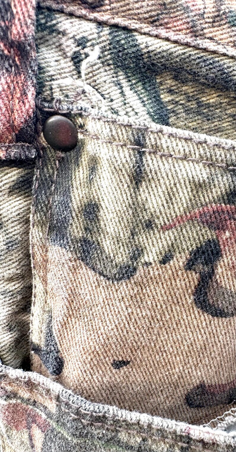 May include: Close-up of camouflage-patterned fabric, featuring shades of brown, green, and black. A dark button is visible on the pocket. The fabric appears to be denim or a similar sturdy material, with visible stitching.