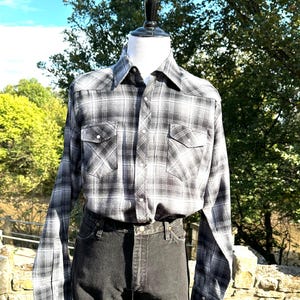 May include: A black and white plaid button-down shirt with two chest pockets and long sleeves. The shirt is paired with black denim jeans. The shirt is displayed on a mannequin outdoors with a natural background.