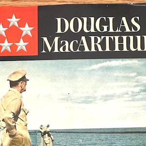 May include: Book cover featuring the title "DOUGLAS MacArthur" in white letters on a black background. A red square with five white stars is in the upper left corner. The image below shows a man in uniform looking out at the sea.