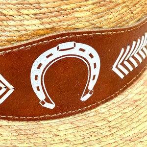 May include: Close-up of a straw hat with a brown leather band. The band features white horseshoe and feather designs. The hat has a woven texture.