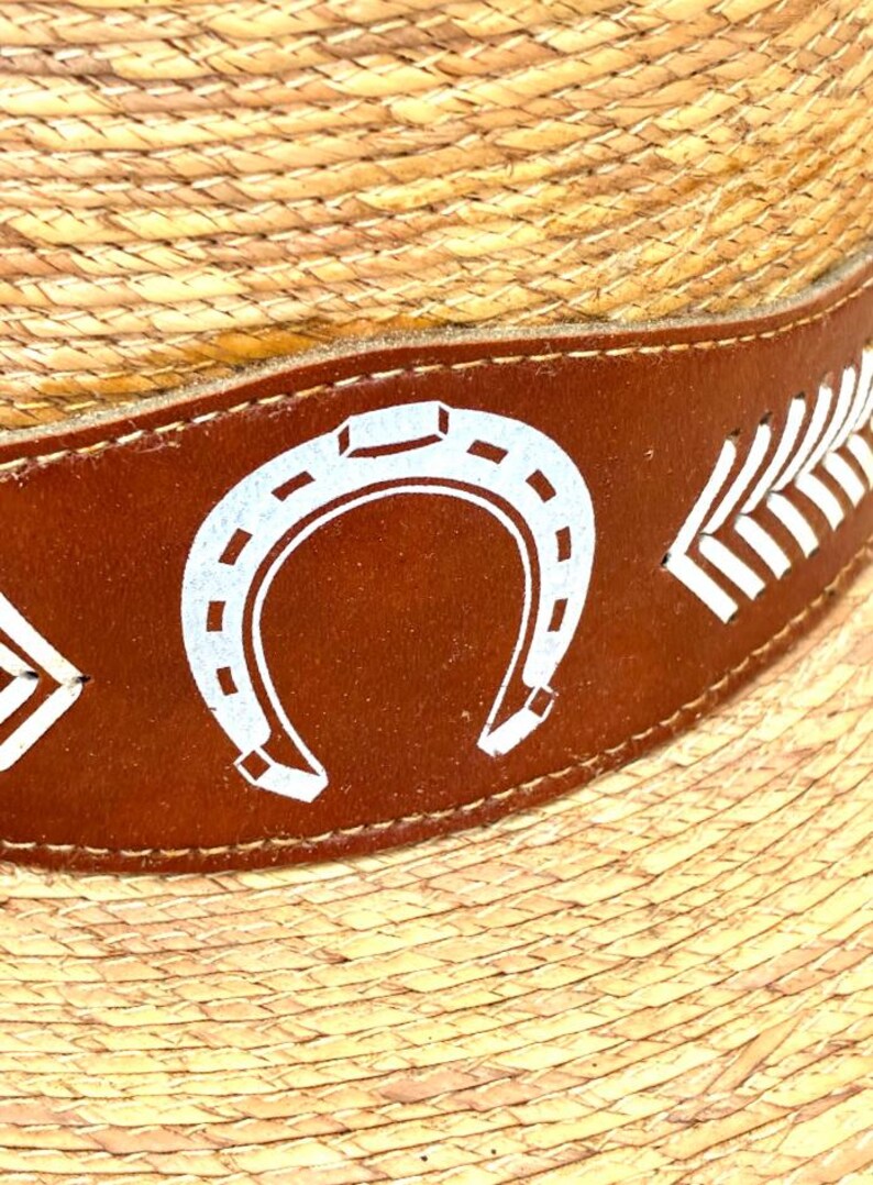 May include: Close-up of a straw hat with a brown leather band. The band features white horseshoe and feather designs. The hat has a woven texture.
