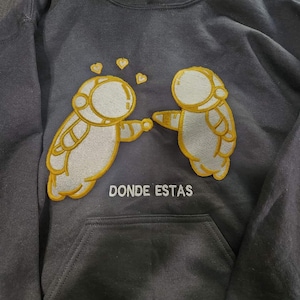 May include: A black hoodie with a white and yellow embroidered design of two astronauts holding hands. The text "DONDE ESTAS" is embroidered below the astronauts.