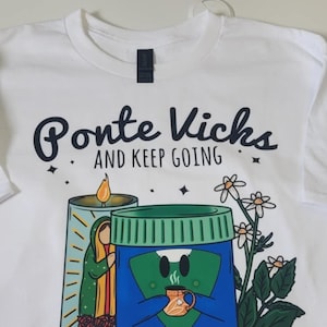 May include: White t-shirt with a black graphic print that says "Ponte Vichs and Keep Going". The graphic features a candle with a religious figure and a blue jar with a face holding a cup of coffee.