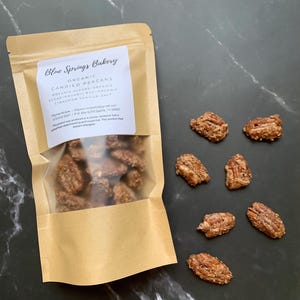 May include: A clear bag of candied pecans from Blue Springs Bakery, with a label that reads "Organic Candied Pecans". Several pecans are scattered on a dark surface. The pecans are coated in a brown, sugary glaze.