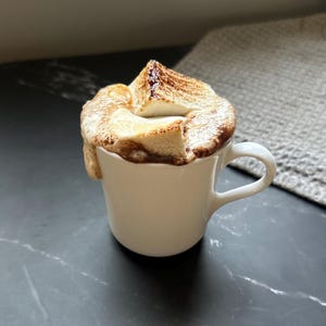 Puede incluir: Una taza de cer&aacute;mica blanca llena de un l&iacute;quido oscuro cubierto con varios malvaviscos grandes y tostados. Los malvaviscos tienen una textura superficial de color marr&oacute;n dorado y carbonizado, desbord&aacute;ndose ligeramente por el borde de la taza.