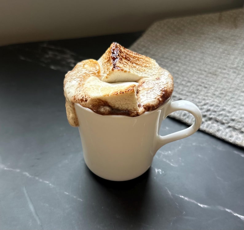 Puede incluir: Una taza de cer&aacute;mica blanca llena de un l&iacute;quido oscuro cubierto con varios malvaviscos grandes y tostados. Los malvaviscos tienen una textura superficial de color marr&oacute;n dorado y carbonizado, desbord&aacute;ndose ligeramente por el borde de la taza.