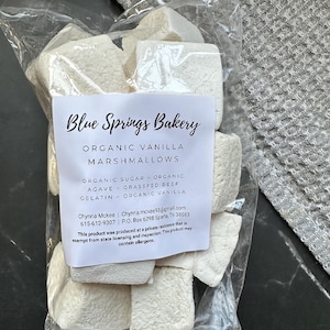 May include: A clear plastic bag filled with white, cube-shaped organic vanilla marshmallows. The white label reads Blue Springs Bakery, Organic Vanilla Marshmallows, Organic Sugar, Organic Agave, Grassfed Beef Gelatin, and Organic Vanilla.