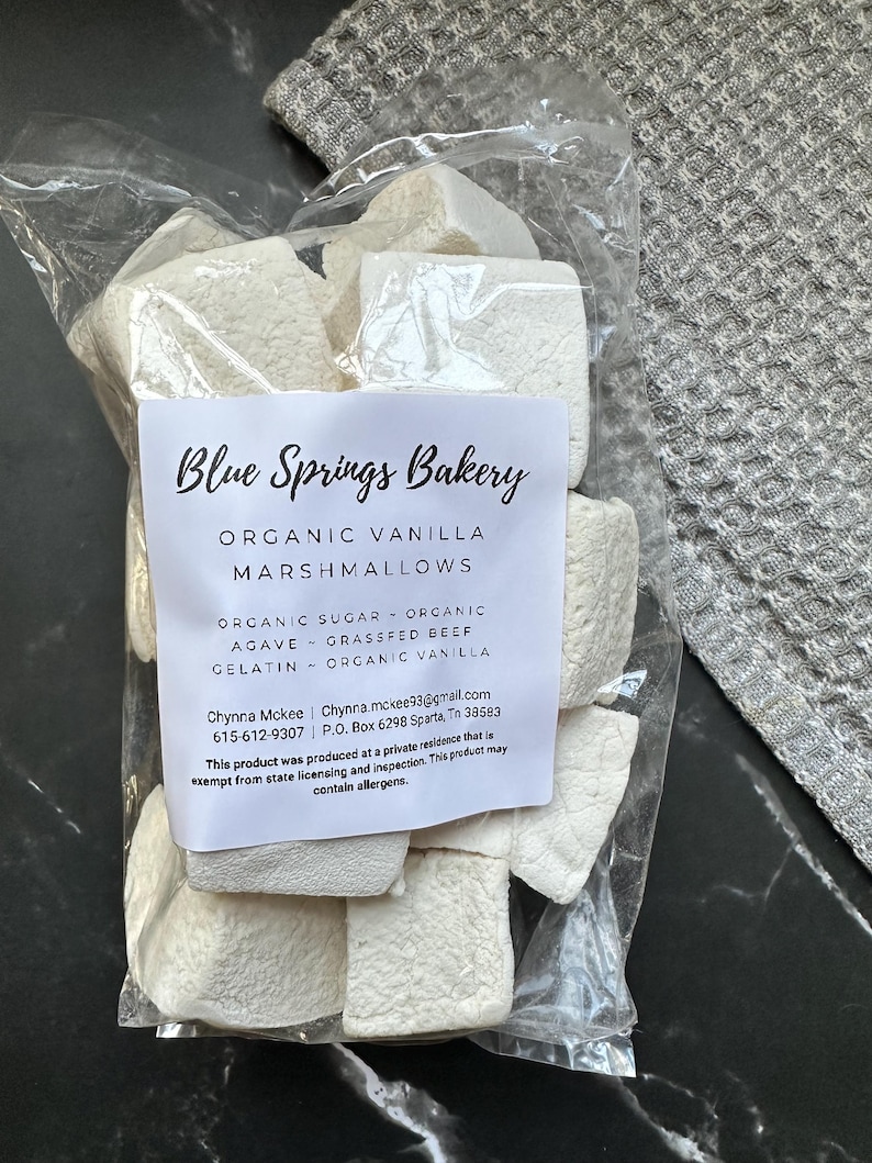 Puede incluir: Una bolsa de pl&aacute;stico transparente llena de malvaviscos blancos c&uacute;bicos de vainilla org&aacute;nica. Una etiqueta blanca en el frente dice Blue Springs Bakery, Organic Vanilla Marshmallows, enumerando ingredientes como az&uacute;car org&aacute;nico, agave, gelatina de res y vainilla.