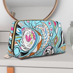 May include: A cosmetic bag featuring a vibrant light blue background with an intricate abstract pattern of swirling black lines, white shapes, and accents in pink, orange, and yellow. The bag has a gold-toned zipper closure with two metal pulls.