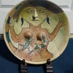 May include: A hand-painted ceramic bowl with a stylized female figure and two lizard-like creatures. The bowl features a light green background, brown and blue accents, and is displayed on a dark wooden stand. The artwork has a folk art style.