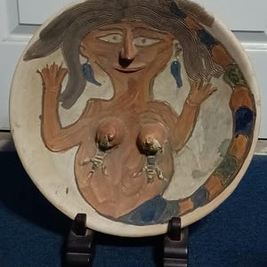 May include: A hand-painted ceramic plate featuring a stylized female figure with long brown hair, blue earrings, and a colorful tail. The figure is painted in shades of brown, blue, and orange. The plate is displayed on a dark brown stand.
