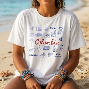 May include: White t-shirt with a blue and red graphic design featuring the word "Colombia" and various illustrations. The design includes a map, a bus, and other cultural symbols. The shirt is worn on a beach.