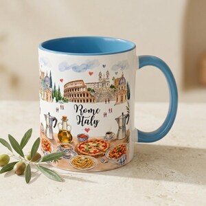 Pode incluir: Caneca de cer&acirc;mica branca com interior e al&ccedil;a azuis, com uma ilustra&ccedil;&atilde;o em aquarela de Roma, It&aacute;lia. O design inclui o Coliseu, outros marcos e comida italiana. A caneca tem as palavras "Rome Italy".