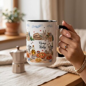 Pode incluir: Uma caneca de cer&acirc;mica branca com interior e al&ccedil;a pretos, com uma ilustra&ccedil;&atilde;o em aquarela de Roma, It&aacute;lia. O design inclui o Coliseu, outros marcos e comida italiana. A caneca tem as palavras "Rome Italy".