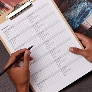 May include: A wooden clipboard holds a password log sheet with the word "PASSWORDS" at the top. The sheet has columns for account/service name, website/app, username, email, and more. A black pen is held over the sheet.