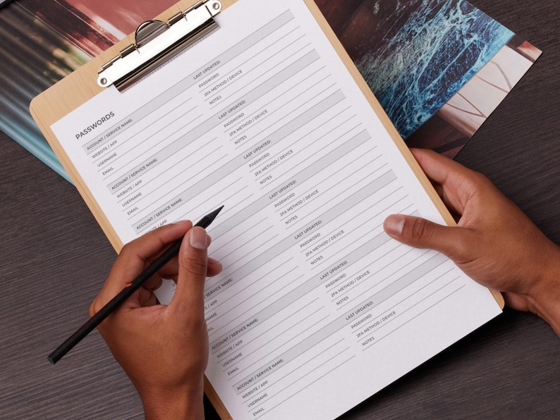 May include: A wooden clipboard holds a password log sheet with the word "PASSWORDS" at the top. The sheet has columns for account/service name, website/app, username, email, and more. A black pen is held over the sheet.