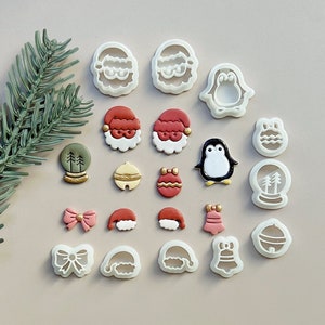 May include: A selection of Christmas cookie cutters featuring festive designs such as Santa faces, penguins, and snow globes. The cutters are white with red, gold, and black details, displayed on a neutral surface with a sprig of evergreen. Metric measurements.