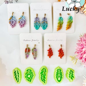 May include: A selection of handmade feather earrings in shades of purple, teal, orange, and red, presented on white cards. The earrings feature gold-coloured posts. Green leaf-shaped cutters are at the bottom. The word "Lucky" is visible.