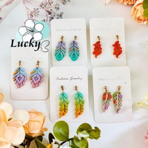 May include: A selection of feather-shaped earrings in shades of blue, orange, purple, and green. Each pair is presented on a white card marked "Fashion Jewelry" with gold-tone earring hooks. The word "Lucky" is visible in the image.